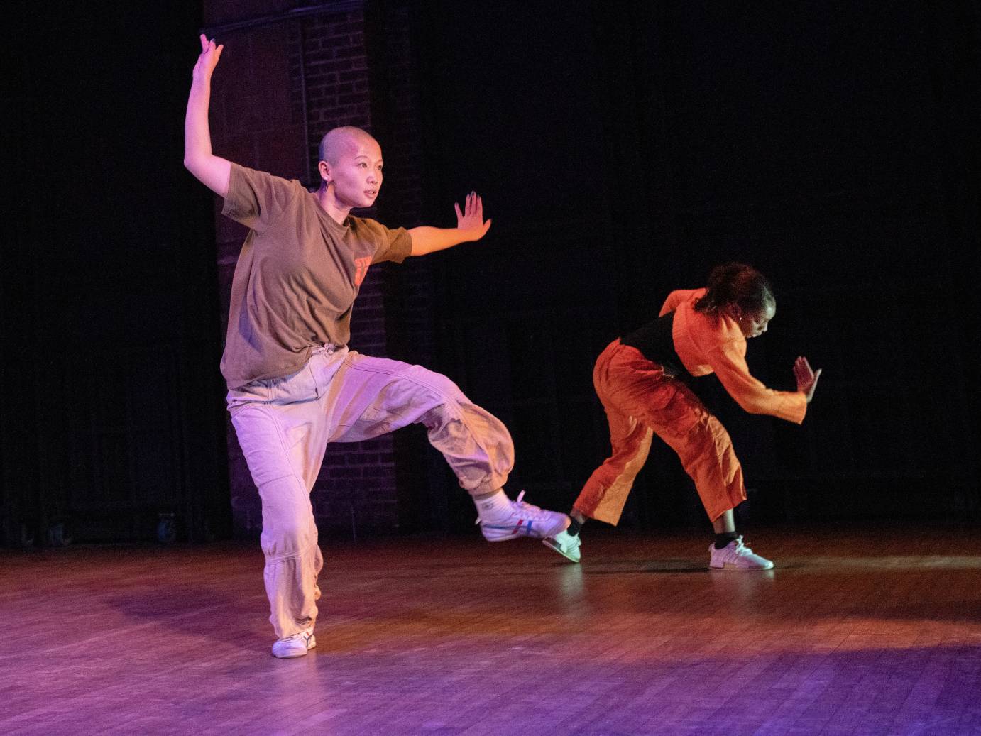 a bald Asian woman in loss fitting khaki pants and a brown t-shirt balances on one leg, while her partner a young Black woman dress in mostly bright orange bends forward..